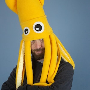 May include: A bright yellow squid hat with large, cartoonish eyes. The hat features long, tentacle-like appendages and is made of soft fabric. The wearer is wearing a dark gray sweatshirt, and the background is a solid blue.