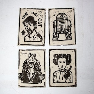 May include: Four black and white linocut prints featuring characters from Star Wars. The prints include Luke Skywalker with the text "ONE WAY OUT", R2-D2, Jar Jar Binks, and Princess Leia holding a blaster. The prints are on a textured, off-white background.