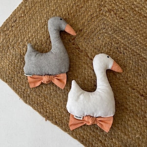 May include: Two plush goose-shaped toys, one in grey and one in white, each featuring a soft orange beak and a matching orange fabric bow tie attached at the base. Both figures have a simple, rounded silhouette and a small white brand tag.