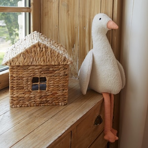 Puede incluir: Una cesta tejida con forma de casa con una pequeña ventana, junto a un ganso blanco de peluche con pico y patas rosas. La casa está hecha de material tejido de color natural. El ganso está sentado sobre una superficie de madera.
