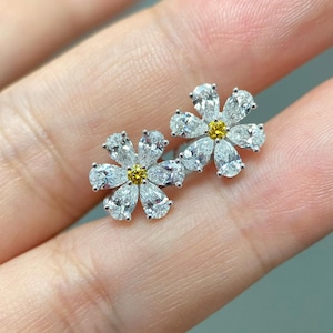 May include: A pair of flower-shaped stud earrings. Each earring has six pear-cut, clear gemstones around a central yellow gemstone, forming a floral pattern. The earrings are set in a silver-coloured metal.