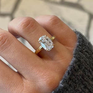 Op de afbeelding: Een gouden ring met een grote, heldere edelsteen in kussenslijpvorm, gezet in een zetting met vier pootjes. De ringband is van glad, gepolijst geelgoud. De edelsteen heeft meerdere facetten die het licht helder en sprankelend weerkaatsen.