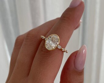 May include: A gold ring featuring a large, oval-cut diamond as the centerpiece. The ring has a delicate band with smaller accent stones on either side of the main diamond. The ring is set in a gold bezel.