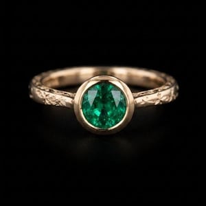 Op de afbeelding: Een gouden ring met een grote, ronde, smaragdgroene edelsteen gezet in een bezel. De band is gegraveerd met een bloemmotief. De ring staat tegen een zwarte achtergrond, waardoor de details en kleur van de ring worden benadrukt.