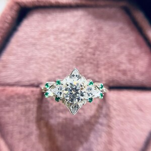 Puede incluir: Un anillo de plata con un gran diamante redondo rodeado de diamantes más pequeños y piedras preciosas verdes. El anillo está en una caja de anillo de terciopelo rosa.