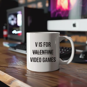 Könnte beinhalten: Weiße Keramiktasse mit dem Text "V IS FOR VALENTINE VIDEO GAMES" in Schwarz. Die Tasse steht auf einer Holzoberfläche mit Tastatur und Computer im Hintergrund. Ein Geschenk für Gamer.