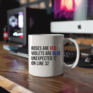 Peut inclure: Mug en céramique blanche avec une anse noire et le texte "Roses are red, violets are blue, unexpected '{' on line 32" en noir, rouge et bleu. Le mug est posé sur une surface en bois.