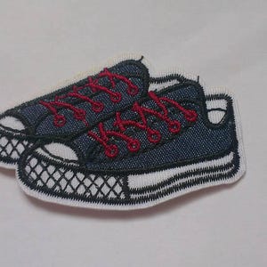 May include: Embroidered patch featuring a pair of navy blue sneakers with red laces. The shoes have white soles and toe caps with a black grid pattern. A fun accessory for clothing or bags.