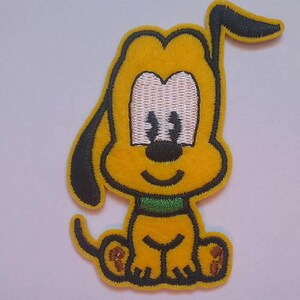 May include: A yellow cartoon dog with black ears and a green collar. The dog is sitting with its legs crossed and has a big smile on its face.