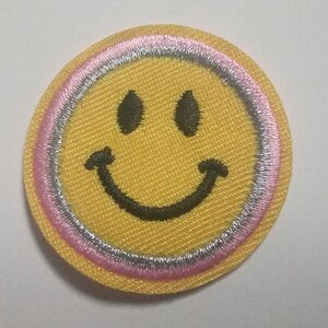Op de afbeelding: Een gele smiley-patch met een roze en zilveren rand. De smiley heeft een zwarte omtrek en een zwarte glimlach.