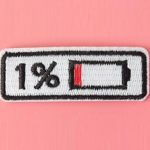 Puede incluir: Parche blanco bordado con contorno negro, que representa un icono de batería con una barra roja que indica un 1% de carga. El texto "1%" también está bordado en negro.