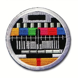 May include: A round embroidered patch with a white border and a black background. The center of the patch features a stylized television test pattern with colored bars and a white grid.