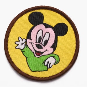 May include: A round, brown-edged patch with a yellow background features a cartoon image of Mickey Mouse wearing a green shirt and smiling.