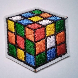 Peut inclure: Écusson brodé représentant un Rubik's Cube. Le cube est composé de carrés aux couleurs vives : rouge, orange, jaune, vert, bleu et blanc, avec des contours noirs. L'écusson a une bordure blanche.