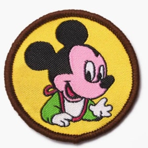 May include: A circular embroidered patch featuring a smiling cartoon mouse with black ears, a pink face, and a green outfit. The mouse is set against a yellow background with a brown border.