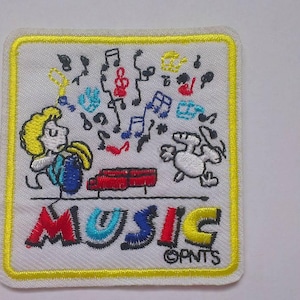 以下が含まれることがあります： 黄色い縁取りのある白い刺繍パッチ。パッチには、楽器を演奏する漫画のキャラクターと音符、犬が描かれています。「MUSIC」という文字はカラフルな文字で書かれており、その下に「GPNTS」とあります。
