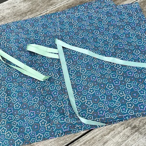 May include: A rectangular fabric piece in a deep teal color featuring a repeating pattern of small, outlined flowers in shades of purple, orange, and white. The fabric is accented with thin, light mint green ribbon ties attached to the top edge.