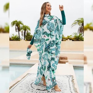 Pode incluir: Um vestido maxi branco com estampa de folhas tropicais verdes. O vestido tem manga comprida e gola alta. A modelo está vestindo o vestido e está em pé em um tapete com padrões brancos e pretos.