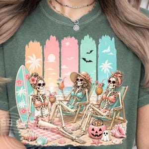 Peut inclure: T-shirt vert avec une scène de plage représentant des squelettes en maillot de bain, lunettes de soleil et chapeaux, se relaxant sur des chaises longues. Une planche de surf, des cocktails et une citrouille d'Halloween sont également présents.
