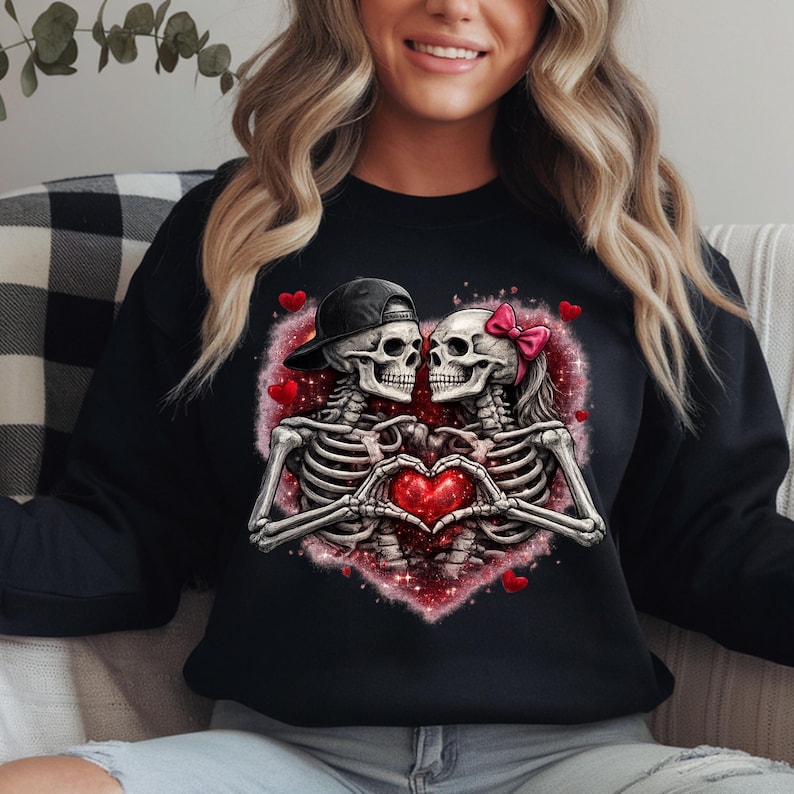 May include: Black sweatshirt featuring a romantic skeleton couple. The skeletons are embracing, forming a heart with their hands. A red heart and pink accents surround them. One skeleton wears a black cap, the other a pink bow.