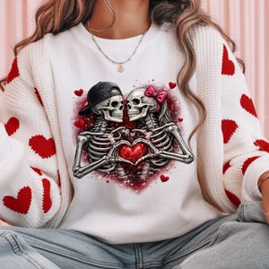 May include: White sweatshirt featuring a graphic of two skeletons forming a heart with their hands. One skeleton wears a black cap, the other a pink bow. Red hearts and a pink glitter background complete the design.