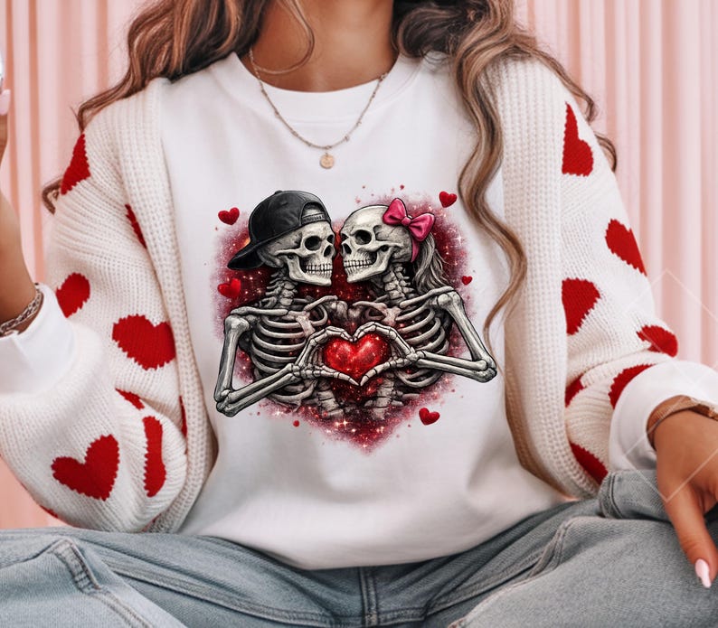 May include: White sweatshirt featuring a graphic of two skeletons forming a heart with their hands. One skeleton wears a black cap, the other a pink bow. Red hearts and a pink glitter background complete the design.