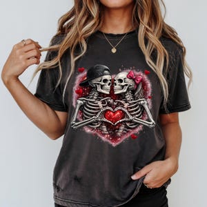 May include: Dark gray t-shirt featuring a romantic skeleton couple inside a heart-shaped design. The skeletons are holding a red heart, with one wearing a black cap and the other a pink bow.