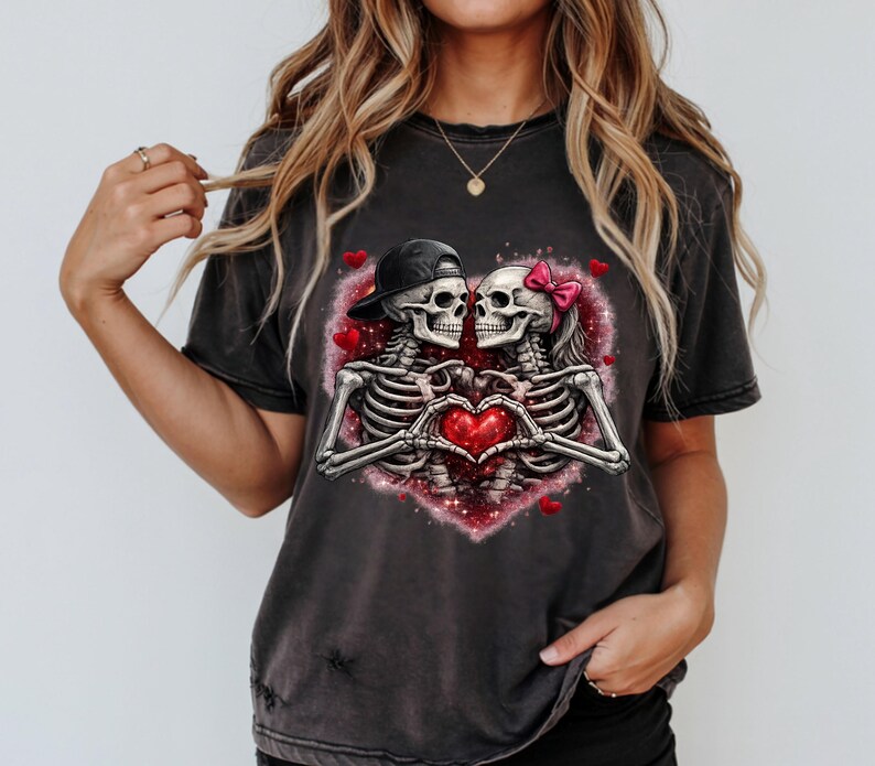 May include: Dark gray t-shirt featuring a romantic skeleton couple inside a heart-shaped design. The skeletons are holding a red heart, with one wearing a black cap and the other a pink bow.