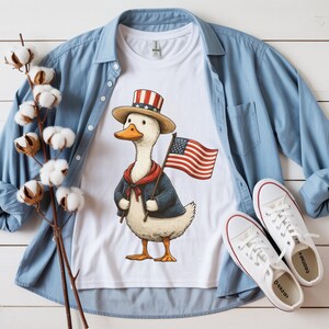 May include: White t-shirt featuring a cartoon duck wearing a patriotic hat and jacket, holding an American flag. The shirt is paired with a blue button-down shirt and white sneakers, creating a casual, all-American theme.