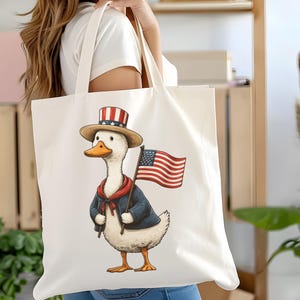 May include: Cream-colored tote bag featuring a patriotic duck illustration. The duck wears a top hat, a blue jacket, and a red scarf, and holds an American flag. The design is printed on a large, rectangular tote bag.