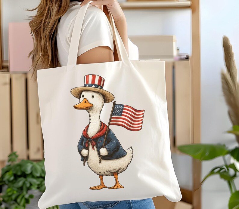 May include: Cream-colored tote bag featuring a patriotic duck illustration. The duck wears a top hat, a blue jacket, and a red scarf, and holds an American flag. The design is printed on a large, rectangular tote bag.