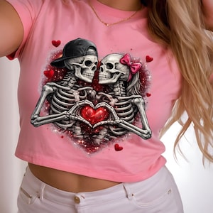 May include: Pink crop top featuring a graphic of two skeletons forming a heart with their hands. One skeleton wears a black cap, the other a pink bow. Red hearts and glittery accents surround the couple.