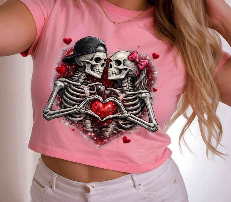 May include: Pink crop top featuring a graphic of two skeletons forming a heart with their hands. One skeleton wears a black cap, the other a pink bow. Red hearts and glittery accents surround the couple.