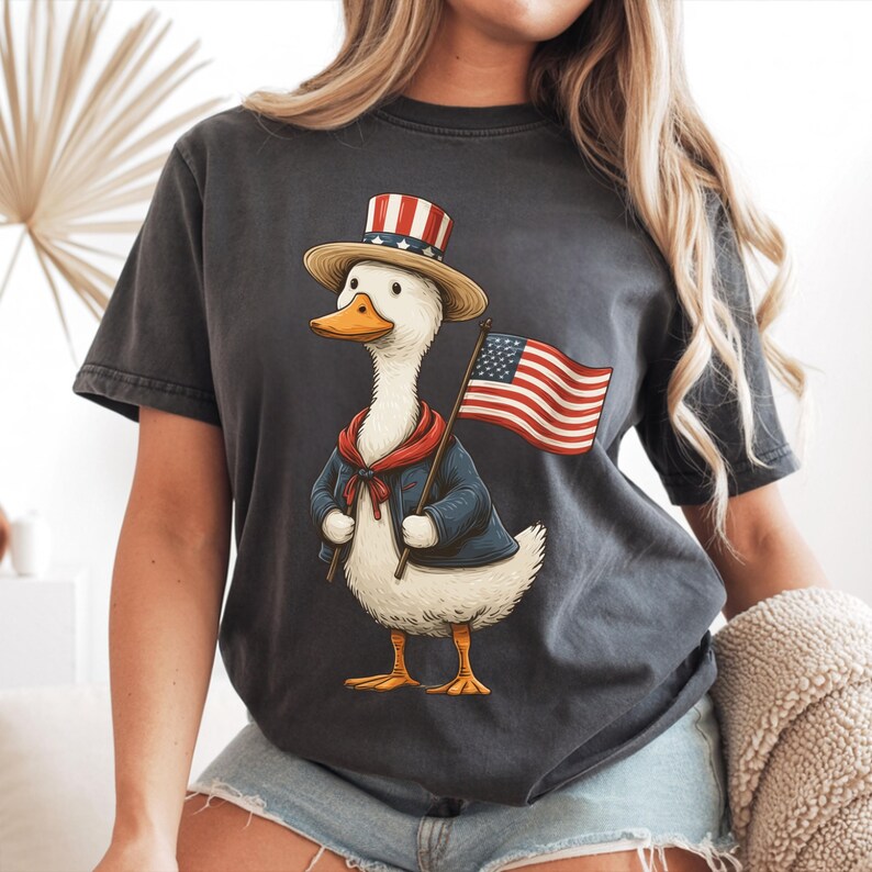 May include: Dark gray t-shirt featuring a cartoon duck dressed in patriotic attire. The duck wears a blue jacket, red scarf, and a hat with red and white stripes, holding an American flag. The design is a whimsical celebration of American pride.