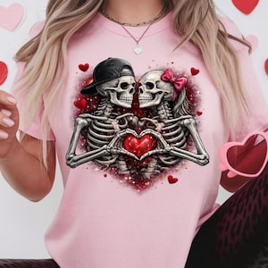 May include: Pink t-shirt featuring a graphic of two skeleton figures embracing, forming a heart with their hands. One skeleton wears a black cap, the other a pink bow. Red hearts and glitter accents complete the design.
