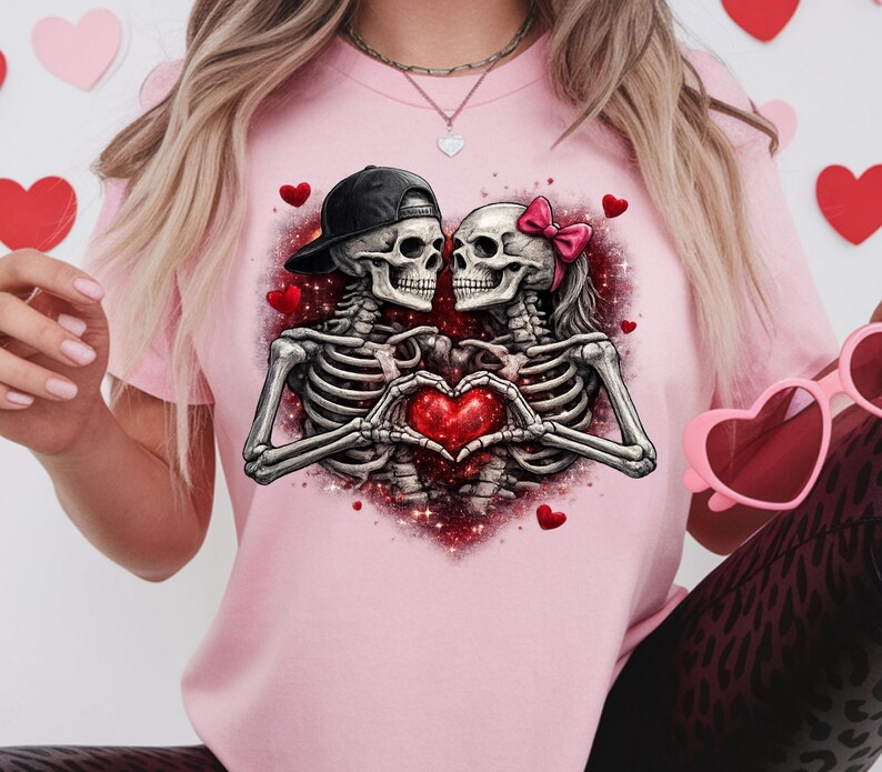 May include: Pink t-shirt featuring a graphic of two skeleton figures embracing, forming a heart with their hands. One skeleton wears a black cap, the other a pink bow. Red hearts and glitter accents complete the design.