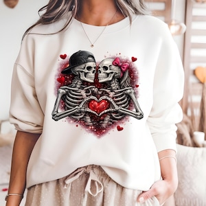 May include: Cream-colored sweatshirt featuring a graphic of two skeletons forming a heart with their hands. One skeleton wears a black cap, the other a pink bow. Red hearts and a red glitter background complete the design.