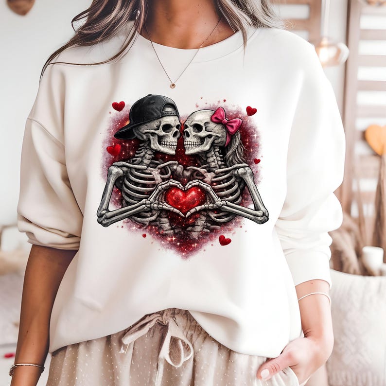 May include: Cream-colored sweatshirt featuring a graphic of two skeletons forming a heart with their hands. One skeleton wears a black cap, the other a pink bow. Red hearts and a red glitter background complete the design.