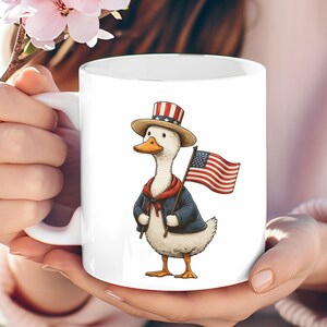 May include: White ceramic mug featuring a cartoon duck dressed in patriotic attire. The duck wears a top hat, a blue jacket, and a red scarf, and holds an American flag. The mug is held by a person.