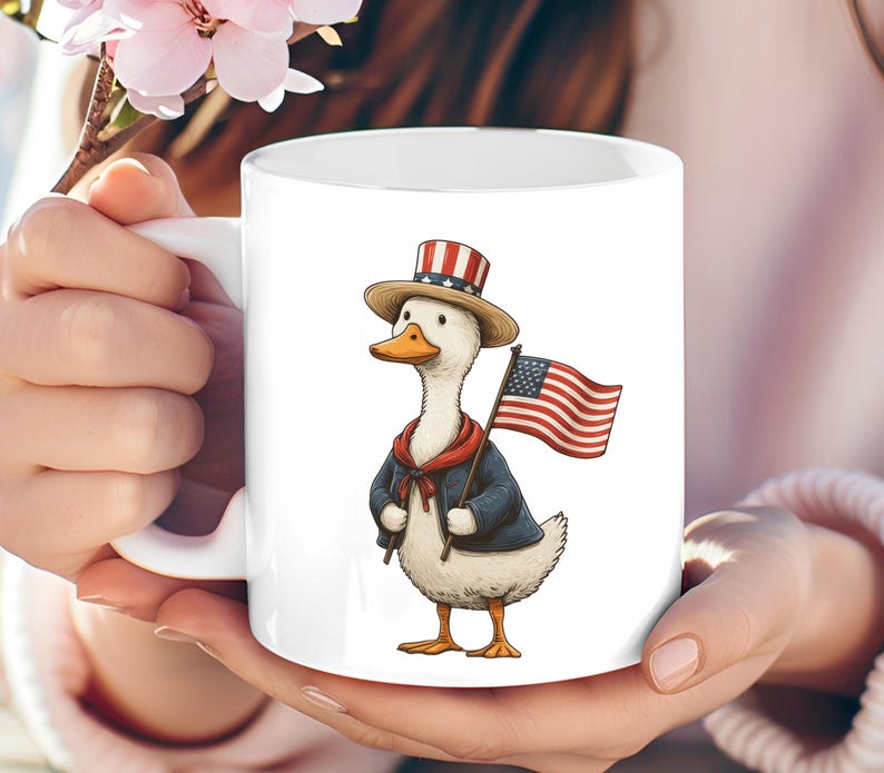 May include: White ceramic mug featuring a cartoon duck dressed in patriotic attire. The duck wears a top hat, a blue jacket, and a red scarf, and holds an American flag. The mug is held by a person.