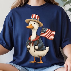 May include: Navy blue t-shirt featuring a cartoon duck dressed in patriotic attire. The duck wears a top hat with red and white stripes, a blue jacket, and a red scarf. It holds a small American flag, perfect for Independence Day celebrations.
