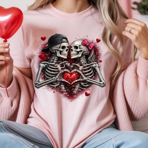 May include: Pink t-shirt featuring a graphic of two skeletons forming a heart with their hands. One skeleton wears a black cap, the other a pink bow. Red hearts and glitter surround the skeletons, creating a romantic theme.