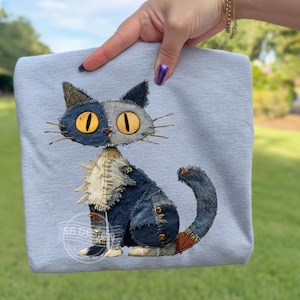 以下が含まれることがあります： パッチワークの猫のデザインが施されたライトグレーのスウェットシャツ。猫はデニムやその他の生地で作られており、黄色い目とひげがあります。猫は座っており、尻尾が丸まっています。デザインには「SB DESIGN」の文字が含まれています。