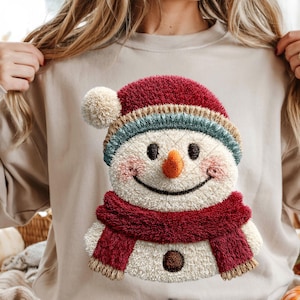 Könnte beinhalten: Beiges Sweatshirt mit einem gestickten Schneemann-Design. Der Schneemann hat einen roten Hut mit einem cremefarbenen Bommel, einen blau-beigen gestreiften Rand, einen roten Schal und eine orangefarbene Karottennase. Das Gesicht des Schneemanns hat einen lächelnden Ausdruck.