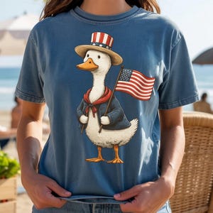 May include: A blue t-shirt featuring a cartoon duck dressed in patriotic attire. The duck wears a hat, jacket, and holds an American flag. The duck has a red scarf and a straw hat with red and white stripes.