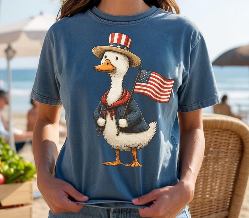 May include: A blue t-shirt featuring a cartoon duck dressed in patriotic attire. The duck wears a hat, jacket, and holds an American flag. The duck has a red scarf and a straw hat with red and white stripes.