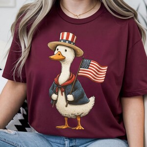 May include: Burgundy t-shirt featuring a cartoon duck dressed in patriotic attire. The duck wears a top hat, a blue jacket, and a red scarf, and holds a waving American flag. The design is a whimsical celebration of American pride.