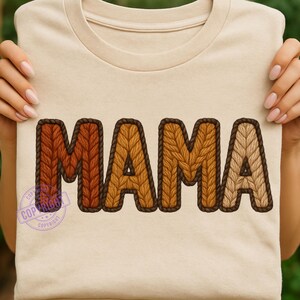 May include: A beige t-shirt featuring the word "MAMA" in large, textured letters. The letters are in shades of brown and tan, with a knitted appearance and a dark brown outline. The shirt is held up by a person.