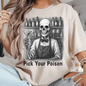 May include: Beige t-shirt featuring a black and white illustration of a skeleton bartender holding a martini glass. The skeleton is behind a bar with bottles and glasses. The text "Pick Your Poison" is below the image.