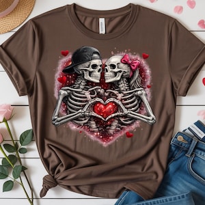 May include: Brown t-shirt featuring a romantic graphic of two skeletons in love, forming a heart with their hands. The skeletons are set against a sparkling heart-shaped background with small red hearts scattered around.